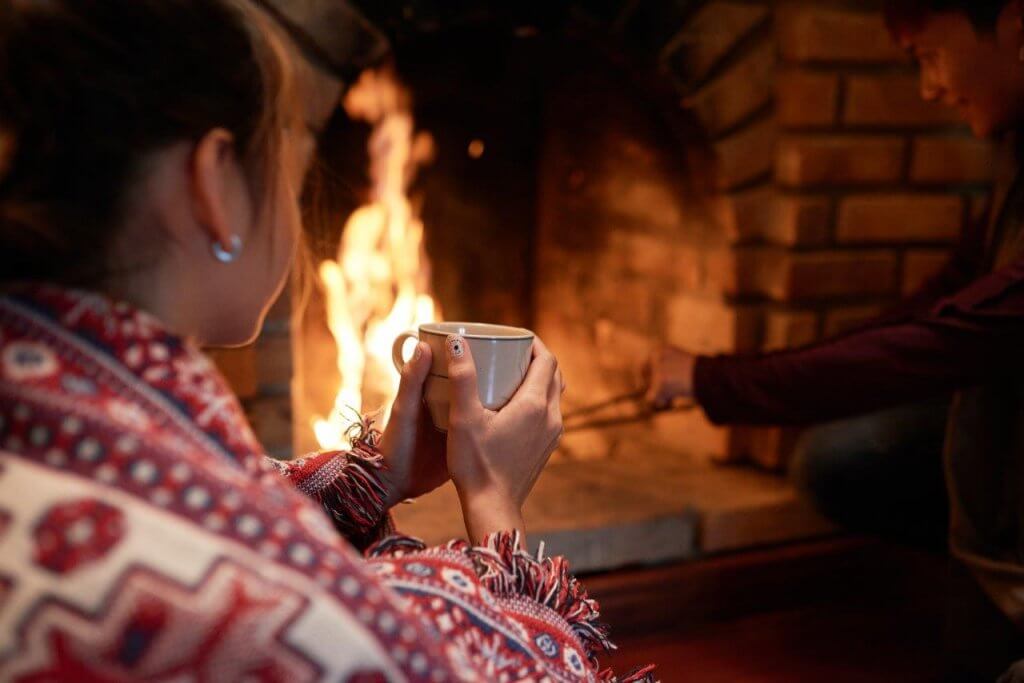 over-the-shoulder-shot-of-woman-warming-her-hands-on-the-mug-of-hot-tea-sitting-at-the-fireplace-her-boyfriend-dealing-with-charcoal