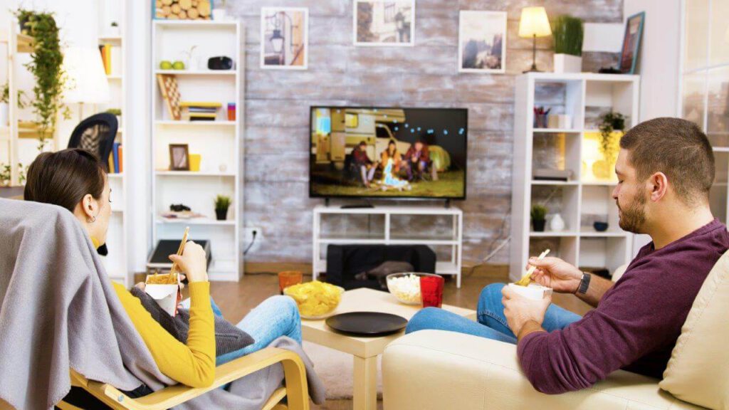 back-view-of-young-couple-sitting-on-comfortable-chairs-eating-noodles-in-front-of-tv-using-chopsticks-cat-sleeping-on-her-cozy-bed-1
